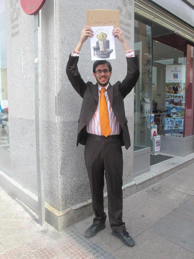 78.-Vestido de traje en una esquina, con un cartel que diga Legen-dario cogido con ambas manos y levantándolo sobre tu cabeza.