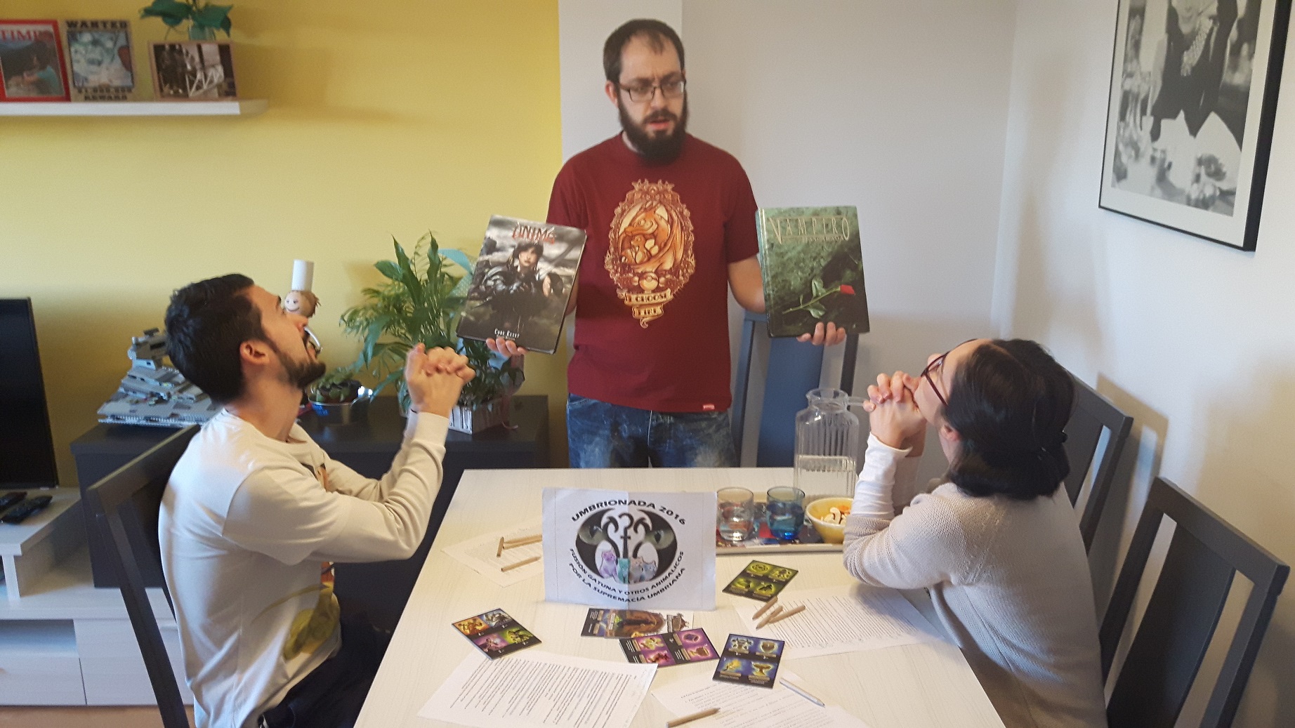 36. En una mesa de juego (rol en mesa) sosteniendo dos manuales como si fueran las tablas de la biblia, mientras los demás jugadores te suplican cleme