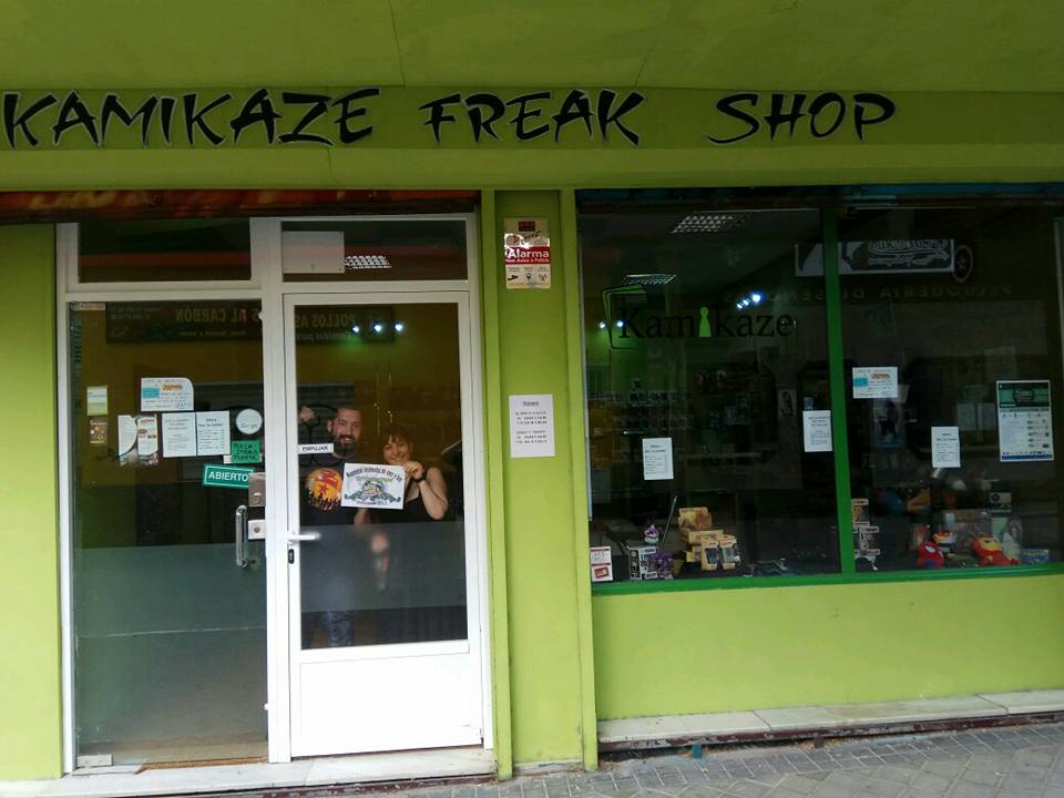 73. En la tienda de Kamikaze, junto al dueño, mientras él y el participante sostienen el logo del equipo.