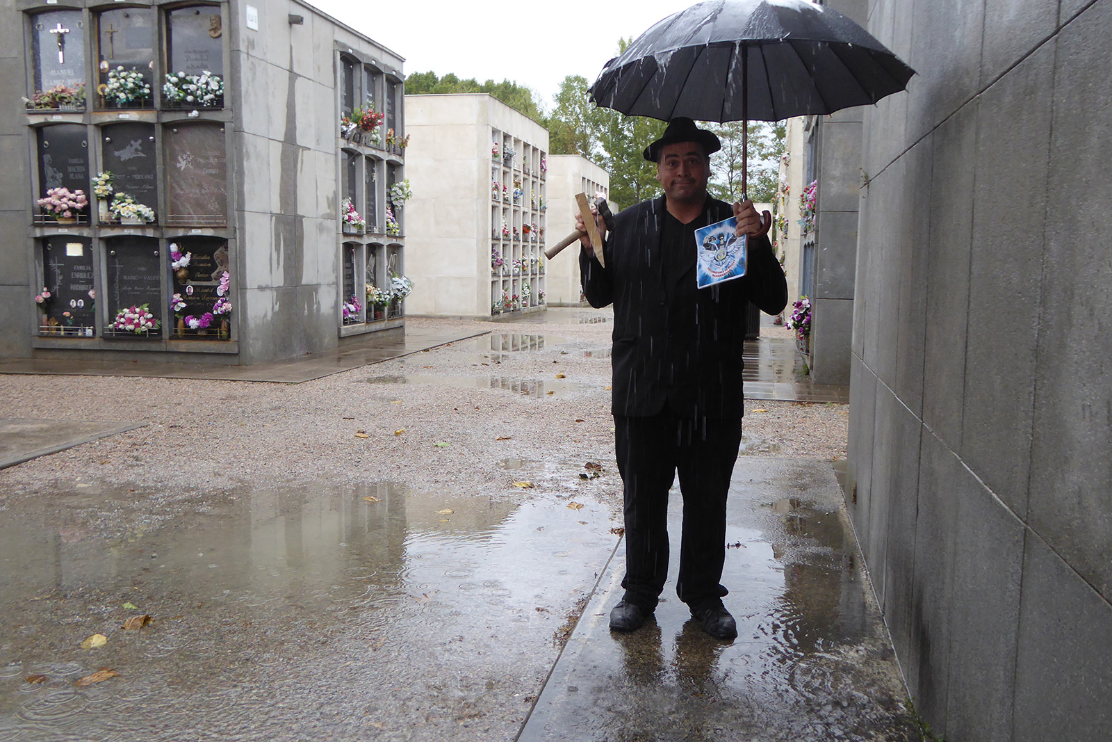94. En un cementerio, vestido de duelo con un paraguas negro mientras llueve.