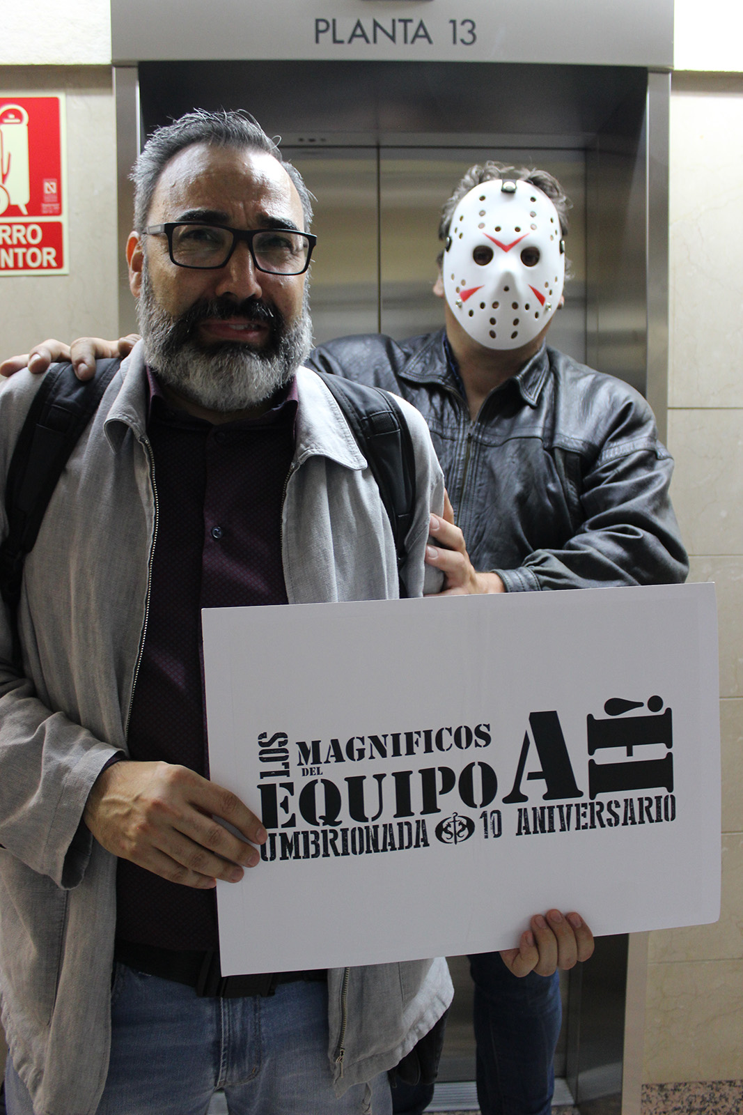 64. Junto al letrero indicativo de la planta 13 de un edificio, con otra persona caracterizada como Jason de Viernes 13 detrás de ti.