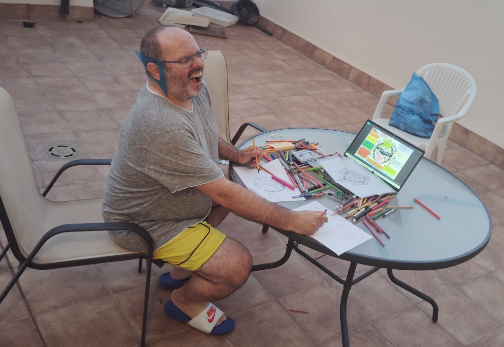 74 - Con un montón de material de artes gráficas, haciendo dibujos, logos y viñetas, con ambas extremidades y cara estresada, mientras en una pantalla