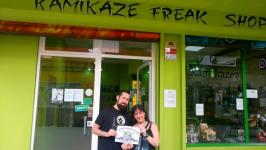 73. En la tienda de Kamikaze, junto al dueño, mientras él y el participante sostienen el logo del equipo.