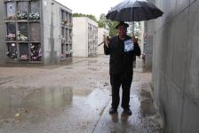 94. En un cementerio, vestido de duelo con un paraguas negro mientras llueve.