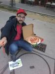 23. Caracterizado como un repartidor junto a una alcantarilla, mientras se sostiene una caja abierta que contenga una pizza completa de pepperoni.